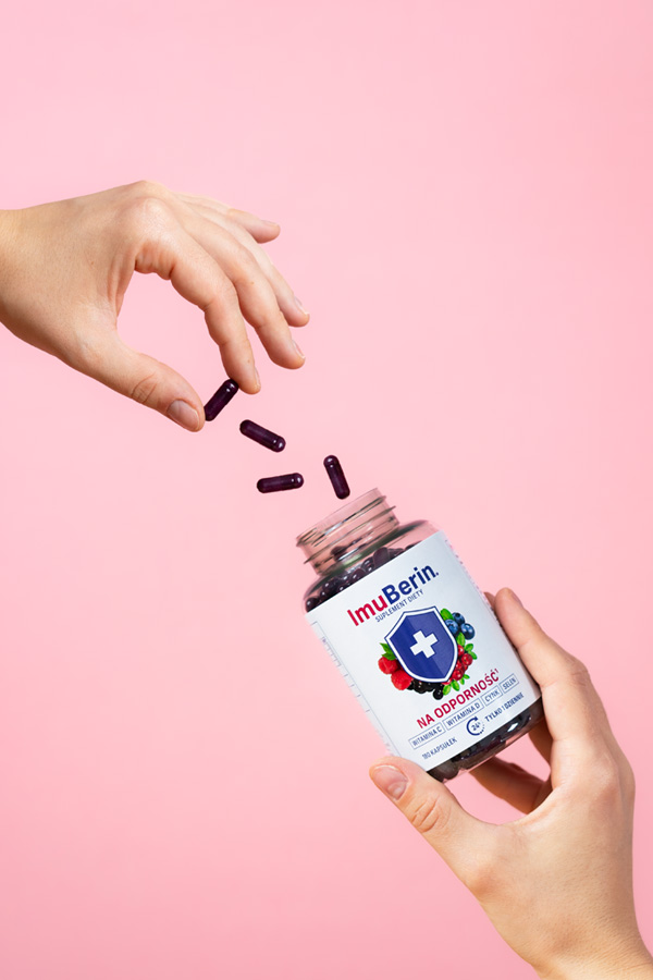 Natural Pharmaceuticals ImuBerin Ranka išpila violetinės kapsulės iš maisto papildo ImuBerin pakuotės ant rožinio fono. Etiketėje nurodyta, kad produktas stiprina imuninę sistemą ir yra vaisių ekstraktų.